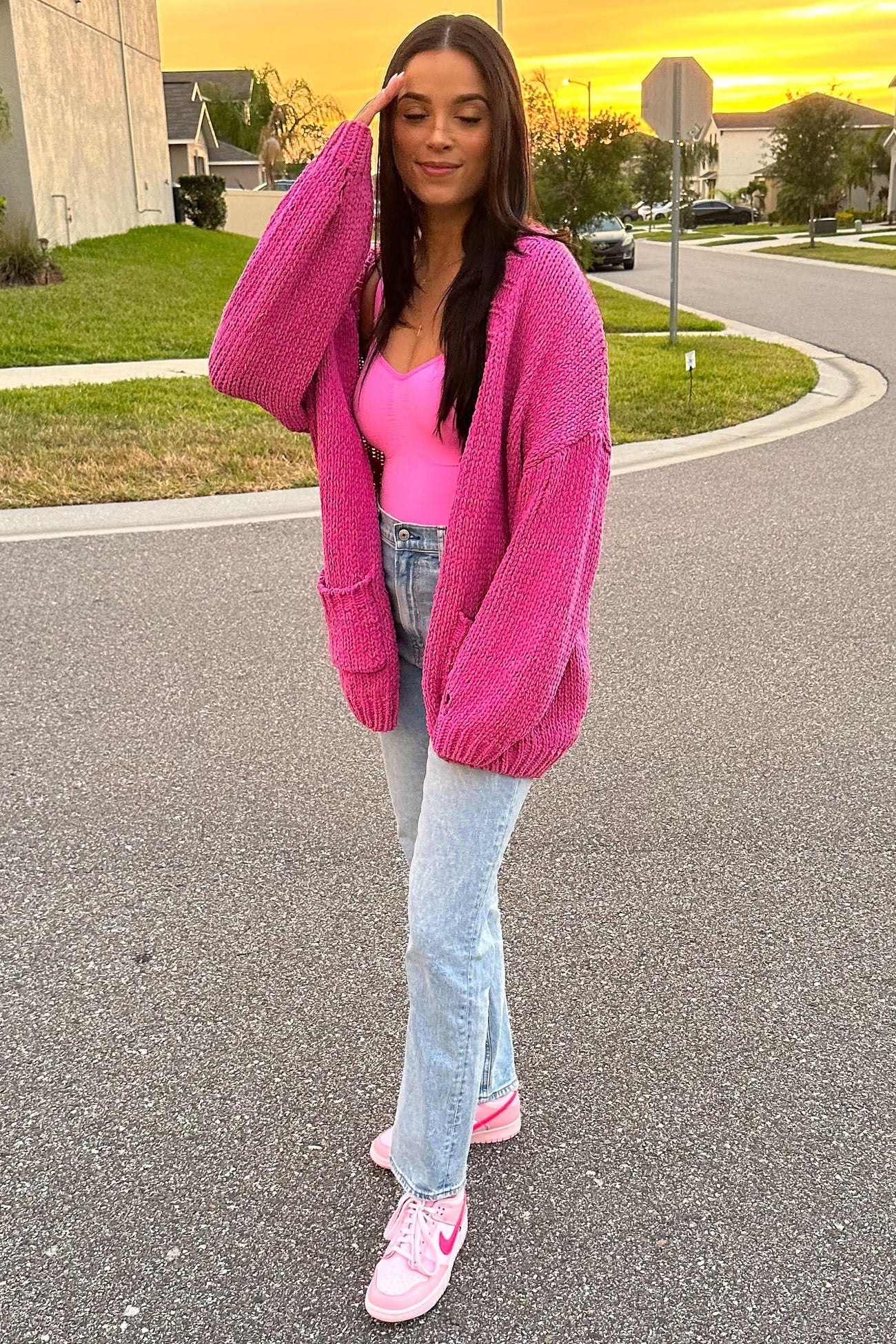 woman wearing a bright pink oversized cardigan, a pink bodysuit, and light blue jeans with pink sneakers, standing outdoors