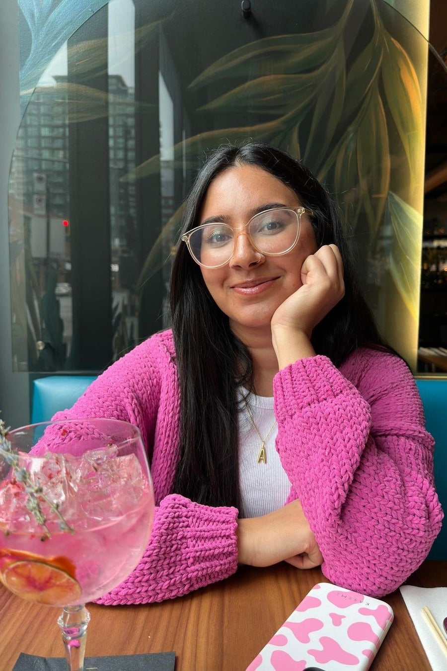 woman wearing a white top and a pink knit cardigan, sitting at a table with a cocktail and phone, smiling