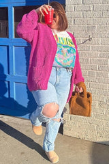 woman wearing a bright pink oversized cardigan, graphic t-shirt, distressed jeans, and beige ankle boots, holding a drink