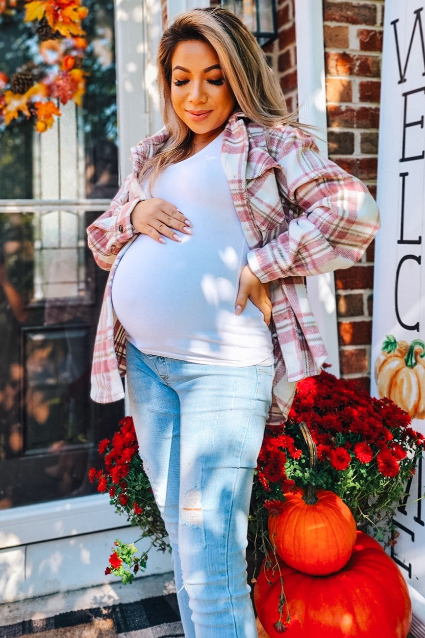 woman wearing a white tank top and a plaid shirt, showcasing her baby bump while standing near flowers and pumpkins