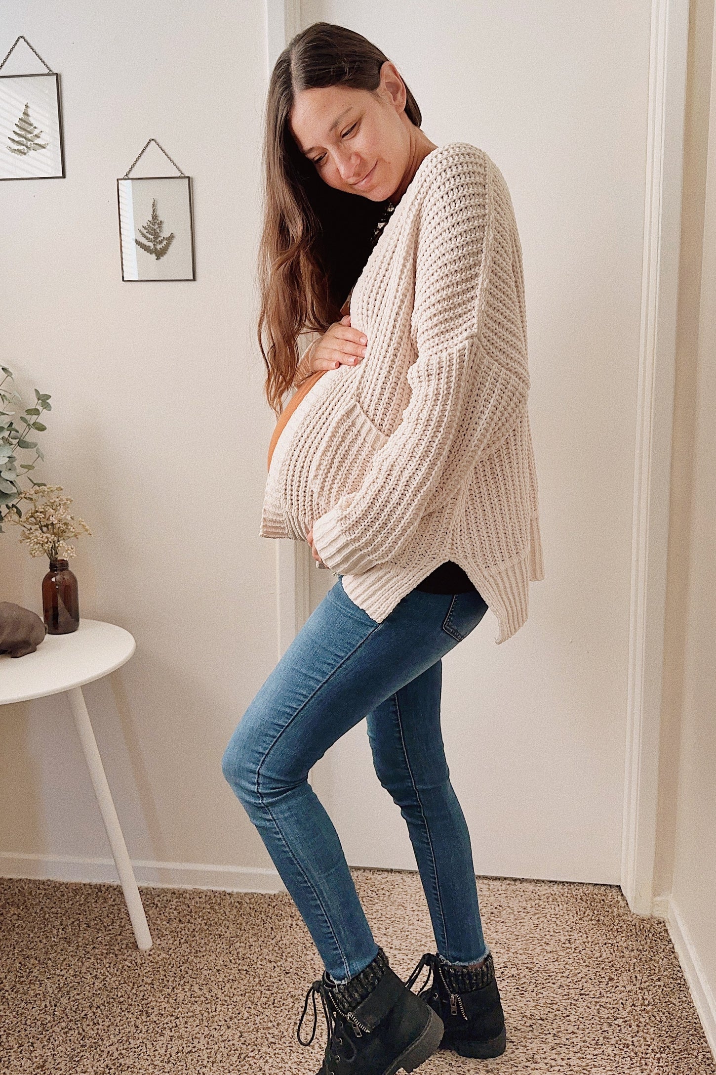 woman wearing a cream chunky knit cardigan and blue skinny jeans, holding her baby bump