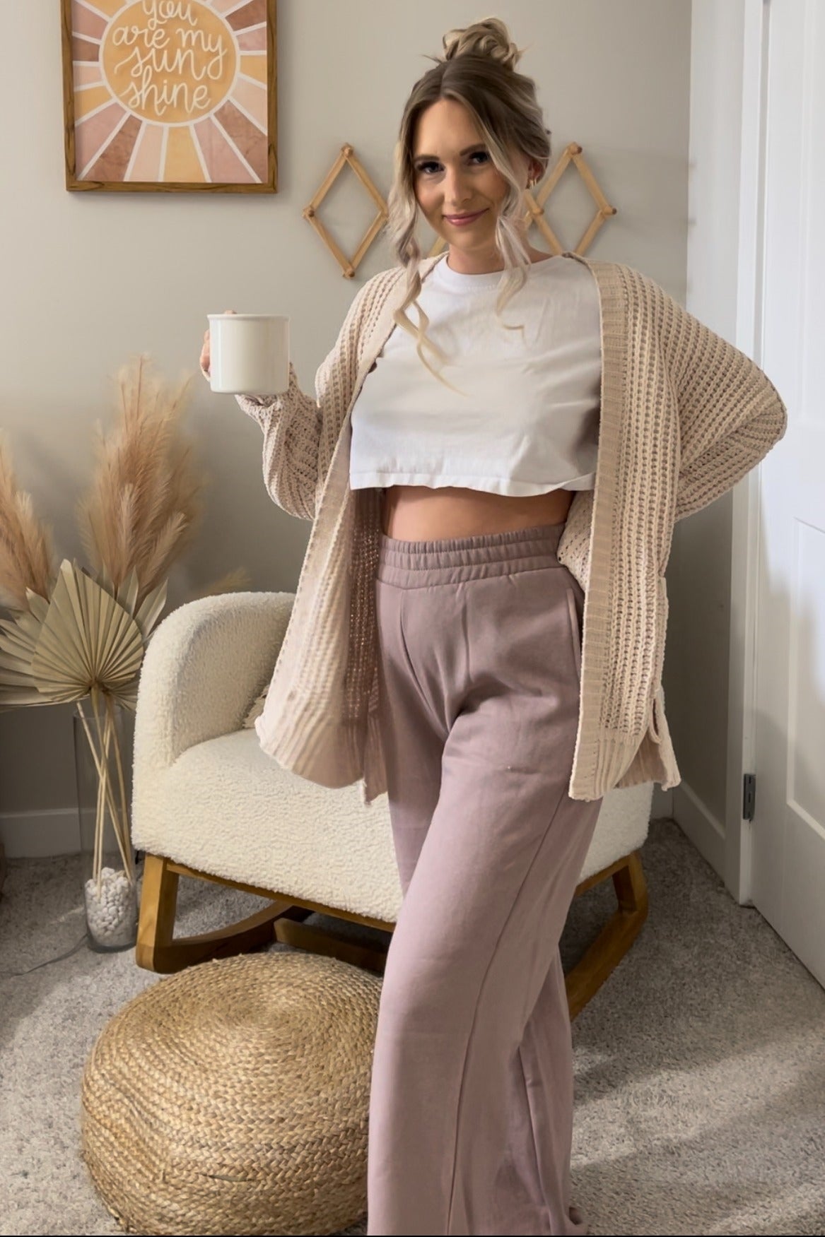 woman wearing a white cropped maternity top, beige cardigan, and light pink maternity pants, holding a mug