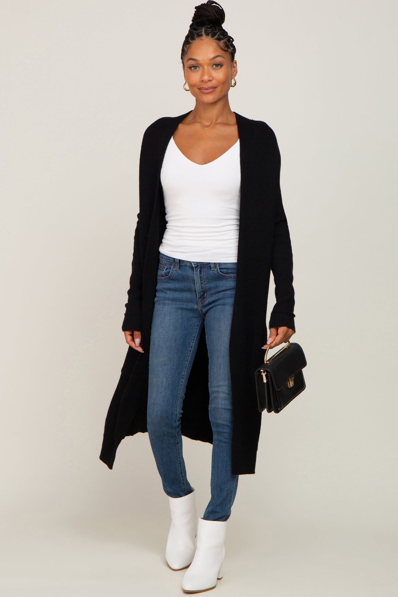 woman wearing a black long cardigan over a white v-neck top with skinny jeans and white ankle boots, holding a black clutch