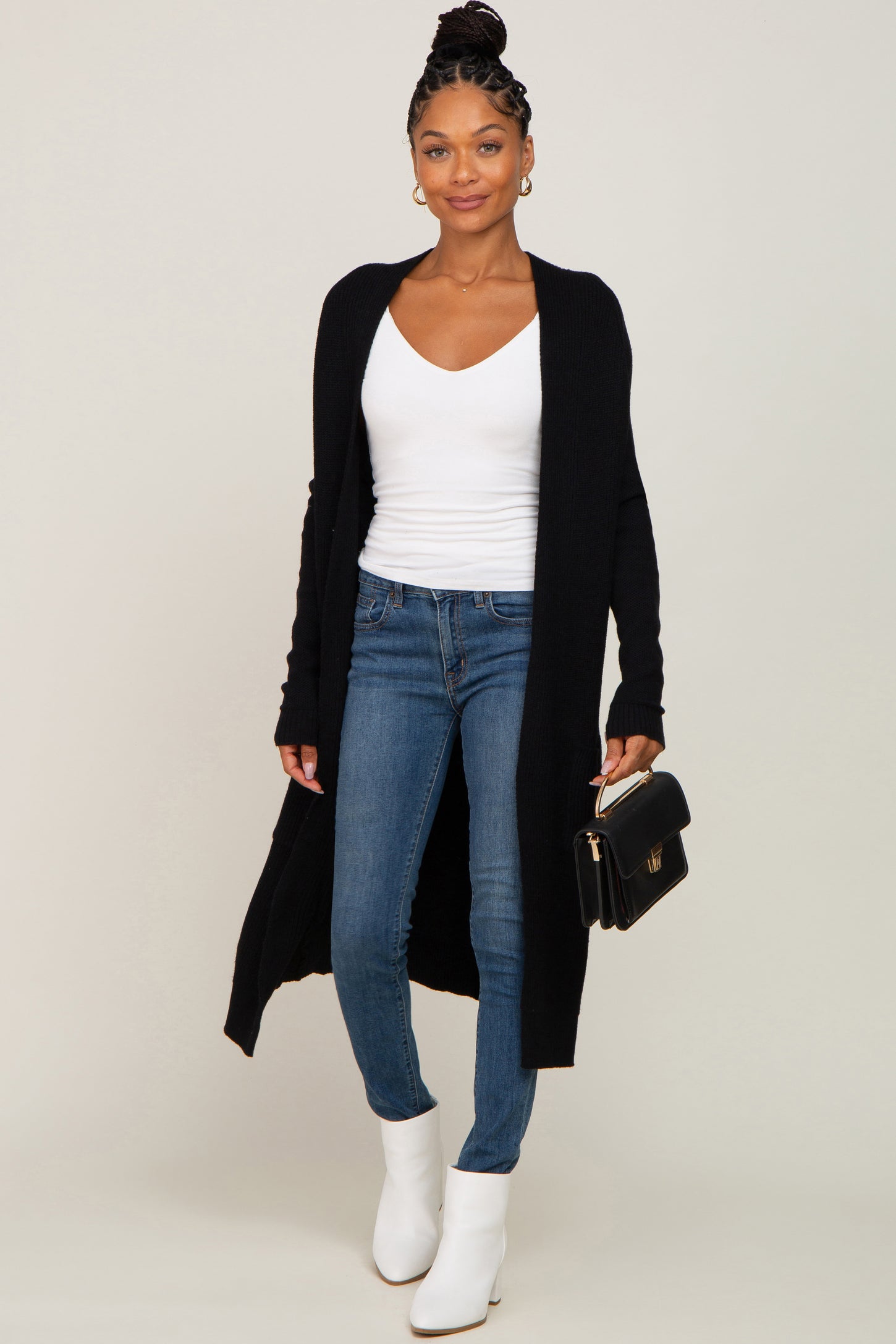 woman wearing a black long cardigan over a white v-neck top with skinny jeans and white ankle boots, holding a black clutch
