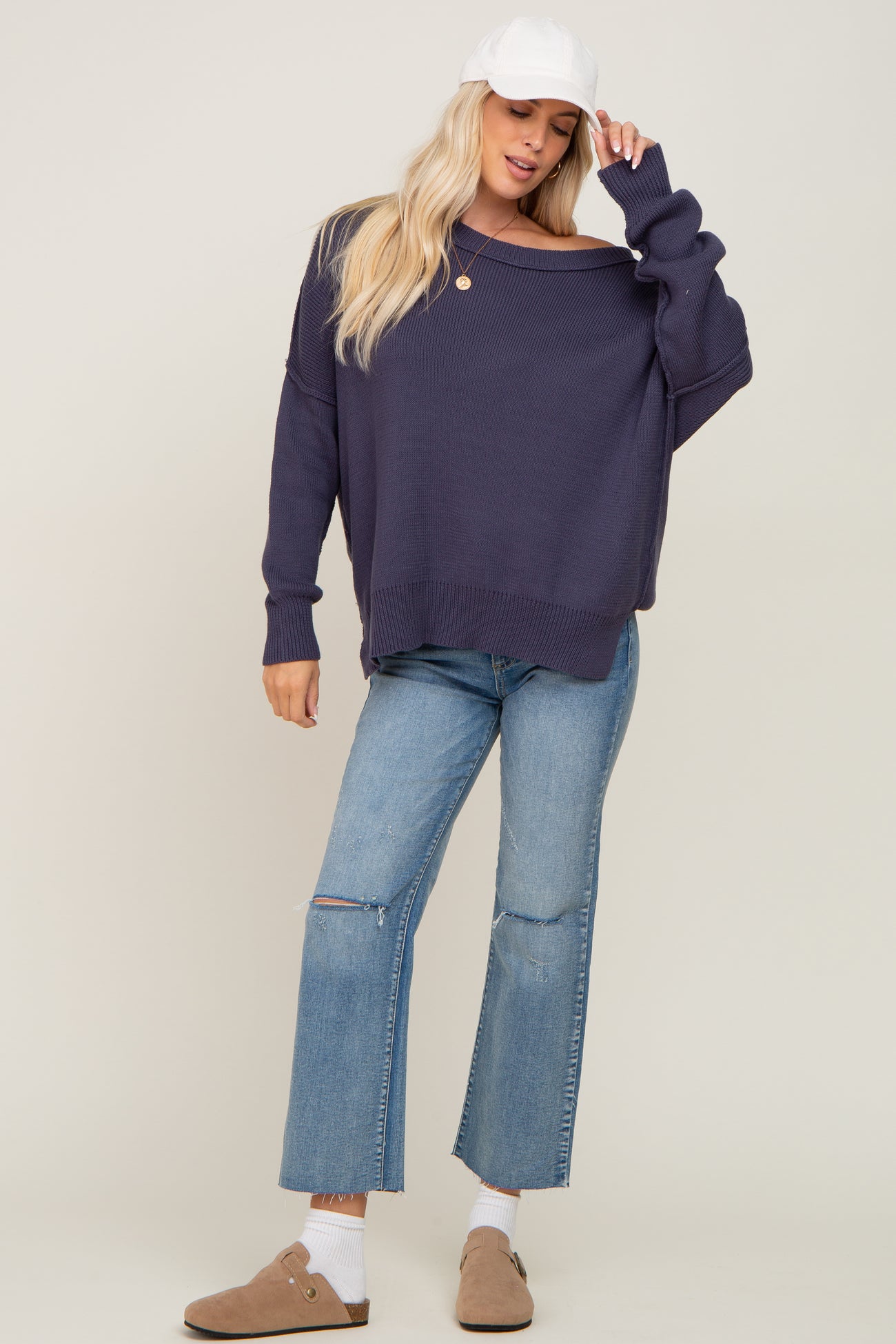 woman wearing a navy oversized sweater with off-the-shoulder detail, paired with light blue ripped straight-leg jeans and a white cap