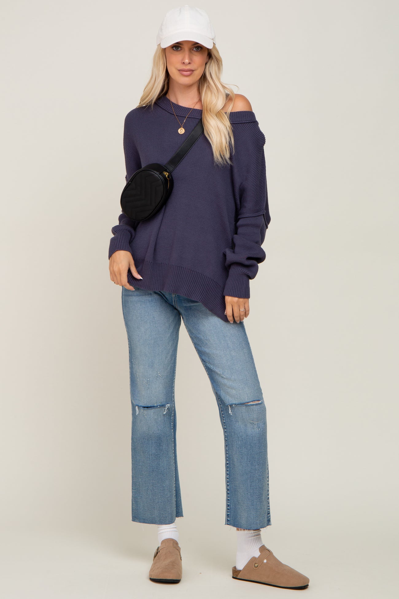 woman wearing a dark purple off-the-shoulder sweater, light blue cropped jeans, and a white cap with a black crossbody bag