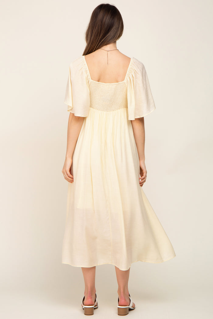 woman wearing a flowy cream maternity dress with short flutter sleeves and a smocked bodice, showing her back
