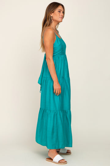 woman wearing a teal maxi dress with a tiered design and spaghetti straps, standing sideways with a relaxed pose