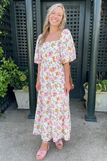 woman wearing a floral print maxi dress with puff sleeves and a square neckline, standing outdoors with plants in the background