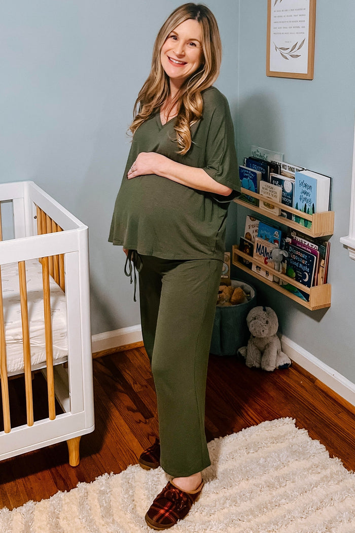 woman wearing a green maternity loungewear set with a tied detail and visible baby bump, standing near a crib