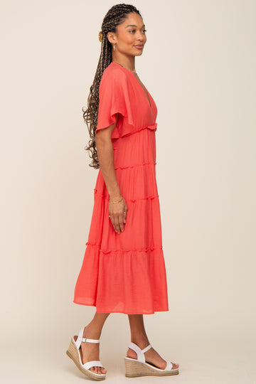 Woman wearing a coral tiered midi dress with flutter sleeves and white wedge sandals against a neutral background