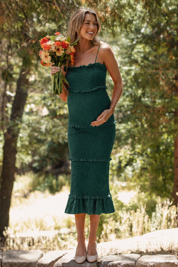 woman wearing a dark green maternity dress with ruffle details, holding a bouquet of flowers and showcasing her baby bump