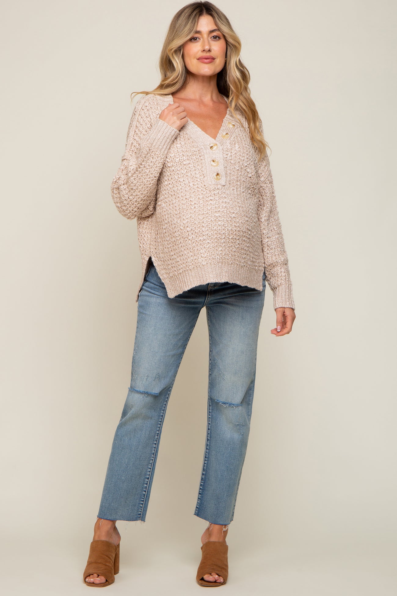 woman wearing a light beige knitted maternity sweater with buttons and paired with light wash jeans, showcasing her baby bump