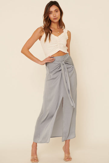 woman wearing a white sleeveless top with ruching and a light grey maxi skirt with a front slit and tie detail