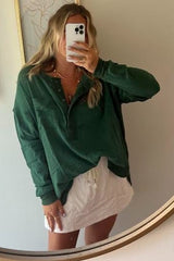 woman wearing a dark green long sleeve button-up top and a white skirt, taking a mirror selfie