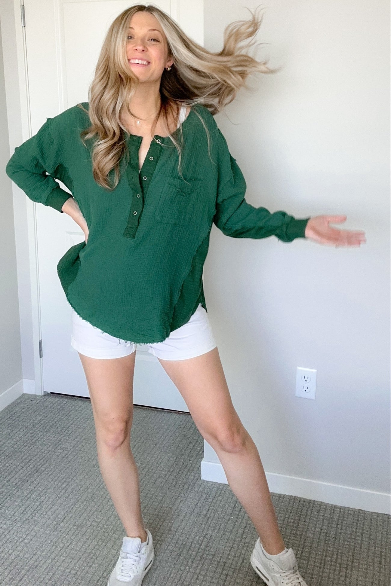 woman wearing a green button-up top and white shorts, smiling with one leg posed and hair flowing