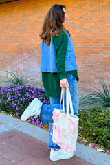 woman wearing a green oversized sweatshirt and denim vest, holding a cream tote bag with pink bows and wearing light blue jeans