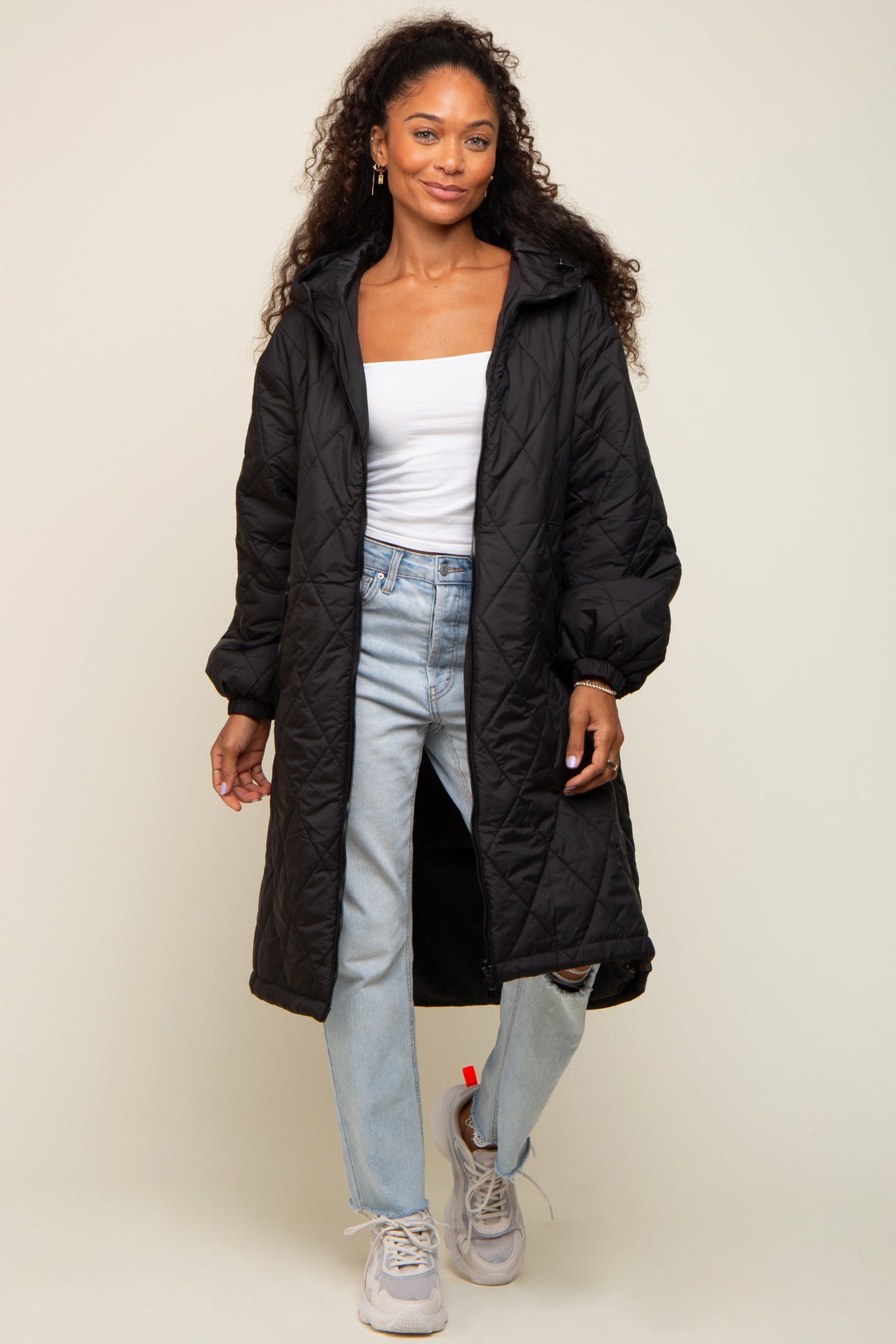 woman wearing a black quilted long coat over a white top and light blue jeans, standing in casual sneakers
