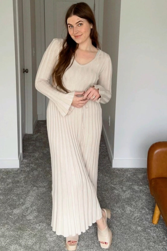 woman wearing a beige long sleeve ribbed maxi dress with flared sleeves and standing on carpeted floor