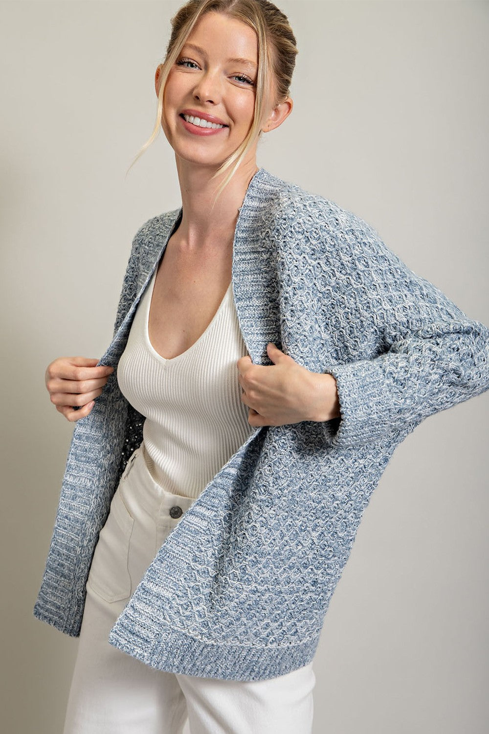 woman wearing a light blue textured cardigan over a white tank top and smiling while holding the cardigan open