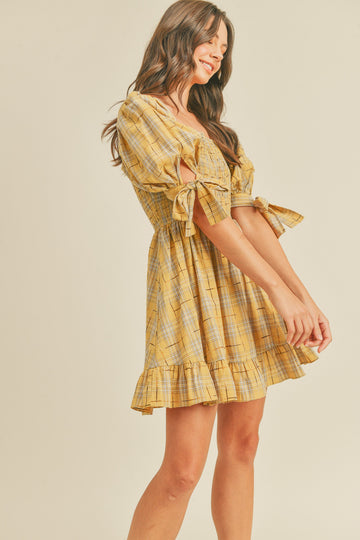 woman wearing a yellow plaid short dress with puff sleeves and tie details, standing sideways and smiling