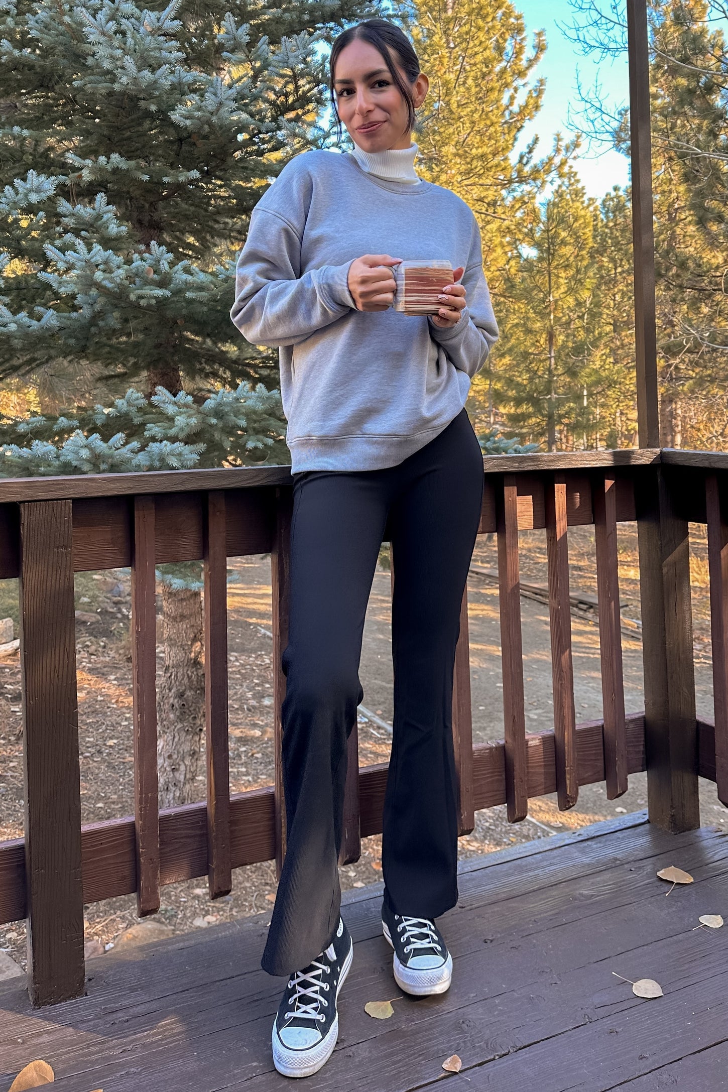 woman wearing a gray sweatshirt and black flared leggings, holding a mug, standing on a deck surrounded by trees