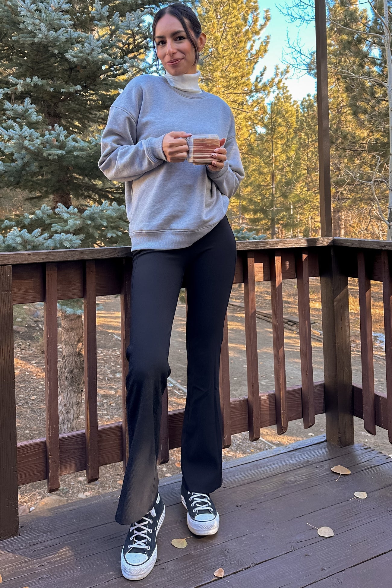 woman wearing a gray sweatshirt and black flared pants, holding a mug while standing on a wooden deck in a forest setting