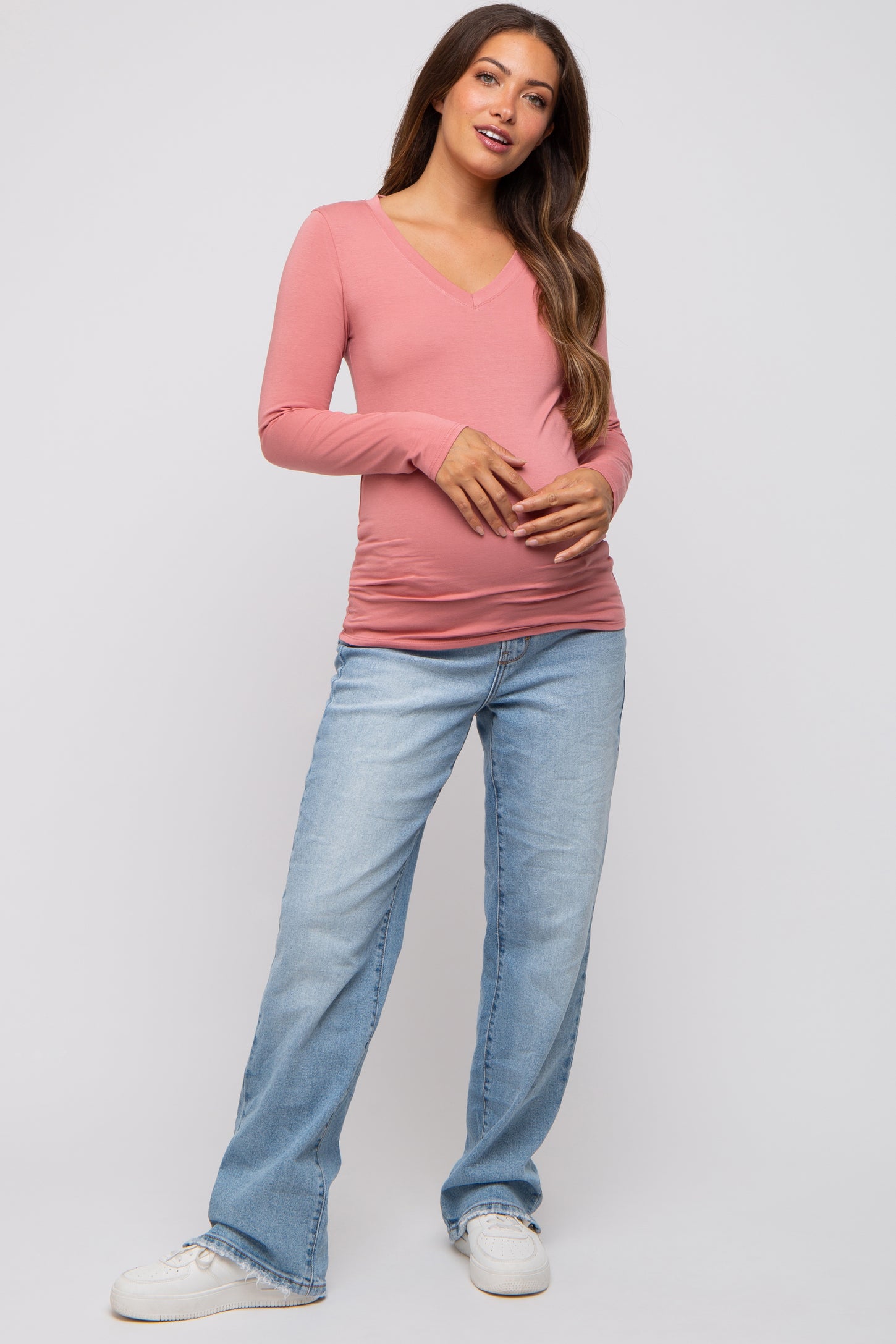 woman wearing a pink long sleeve maternity top and holding her baby bump, paired with light blue jeans