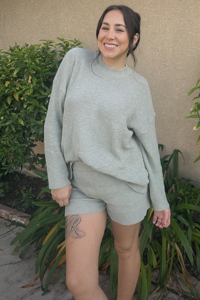 woman wearing a light gray ribbed sweater and matching shorts, smiling outdoors among greenery