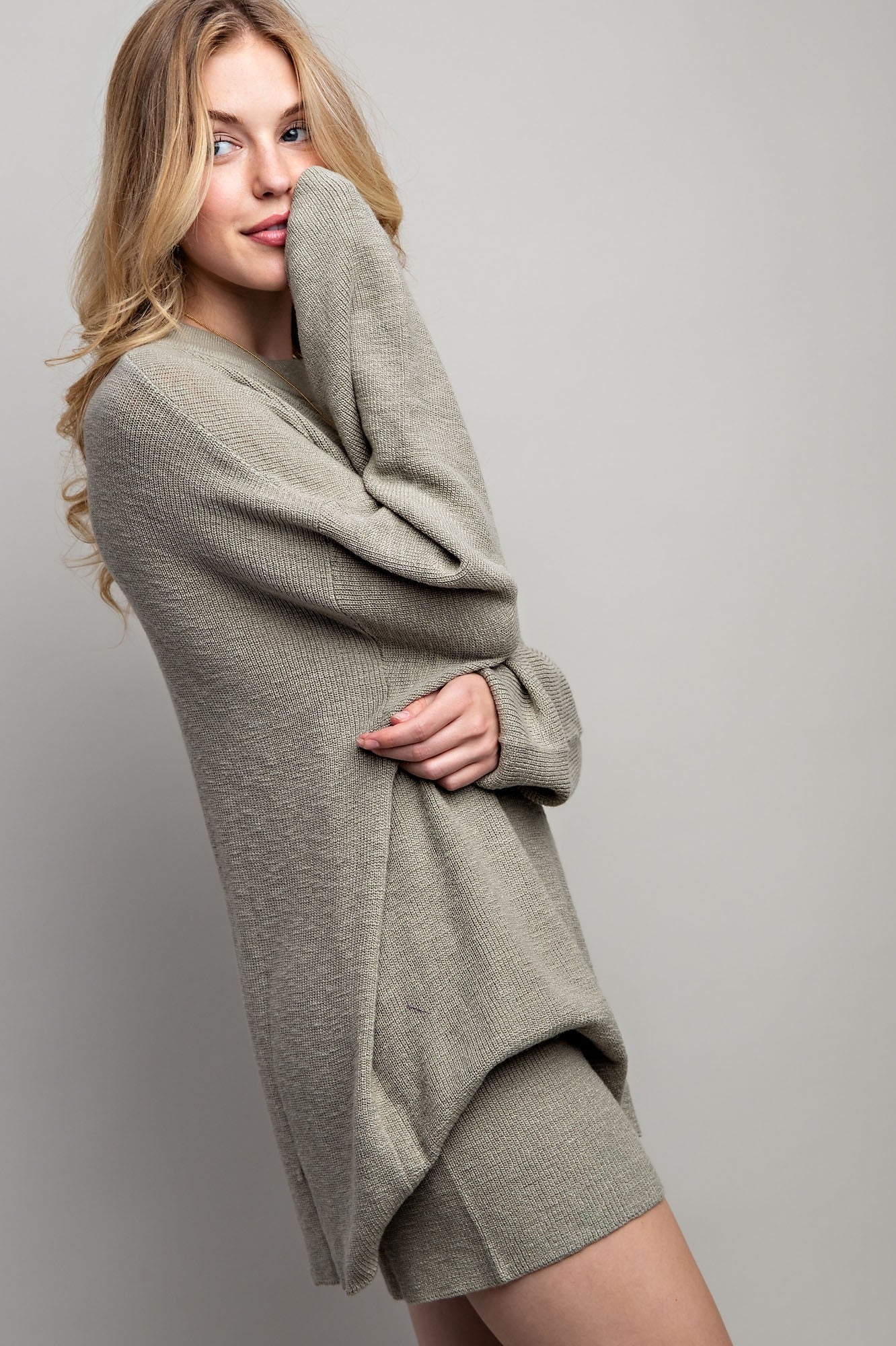 woman wearing a light olive green oversized sweater and matching shorts, smiling and posing sideways