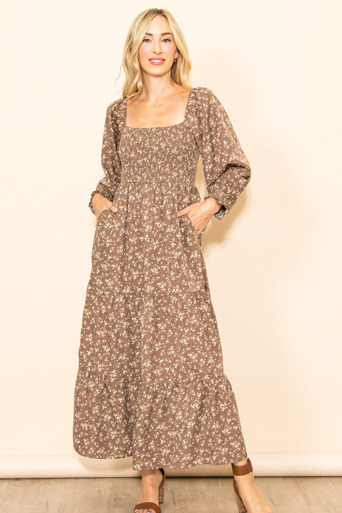woman wearing a brown floral maxi dress with a square neckline and long sleeves, standing with hands in pockets