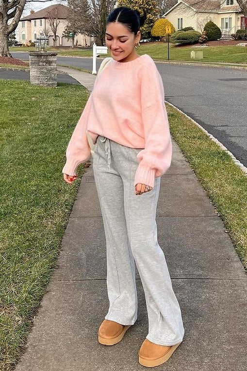 woman wearing a pink oversized sweater with gray flared sweatpants and tan slip-on shoes, standing outdoors on a walkway