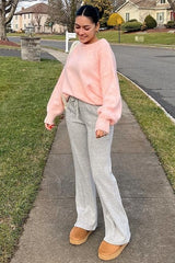 woman wearing a pink oversized sweater with gray flared sweatpants and tan slip-on shoes, standing outdoors on a walkway
