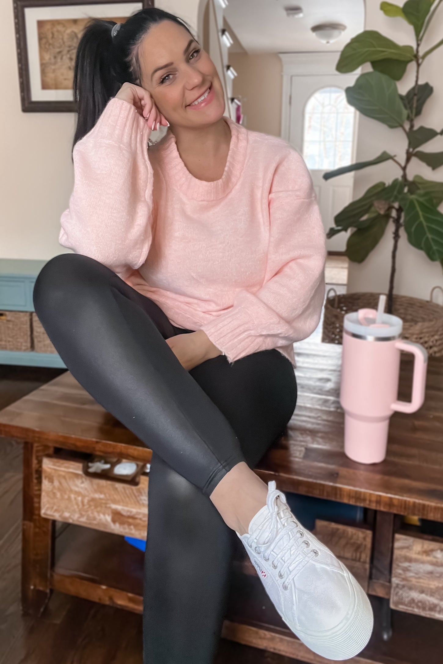woman wearing a light pink oversized sweater and black leggings, sitting on a wooden table with a drink beside her