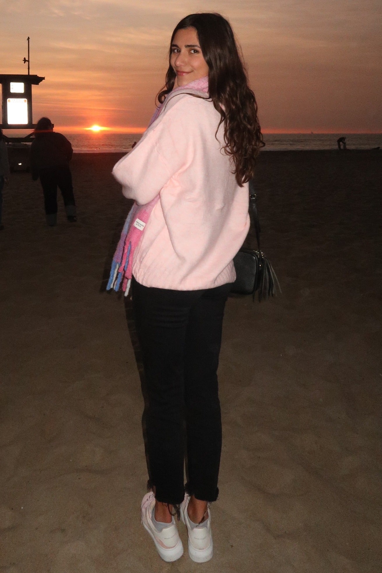 woman wearing a light pink oversized sweater, black jeans, and white sneakers, with a pastel scarf draped around her neck
