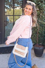 woman wearing a pink long sleeve sweater and blue high-waisted jeans, holding a cream-colored shoulder bag with a chain strap