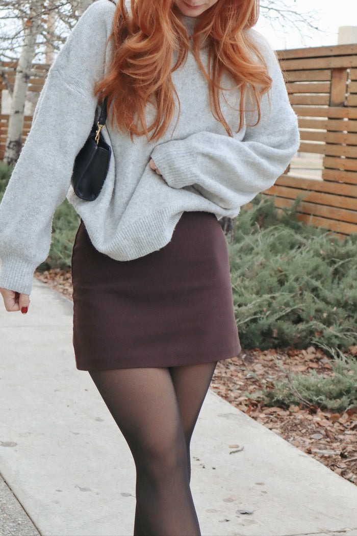 woman wearing a light gray oversized sweater and a brown mini skirt, walking in an outdoor setting