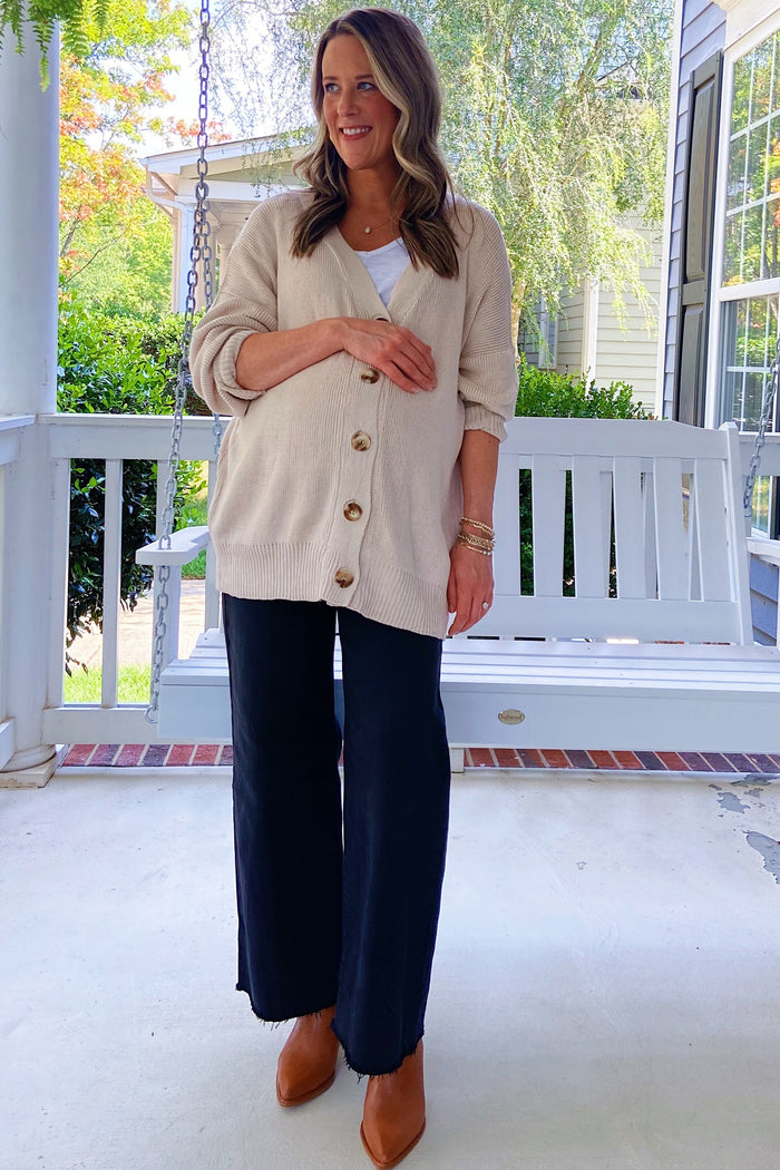 woman wearing a beige cardigan and a white tank top paired with black wide-leg pants, standing by a porch swing