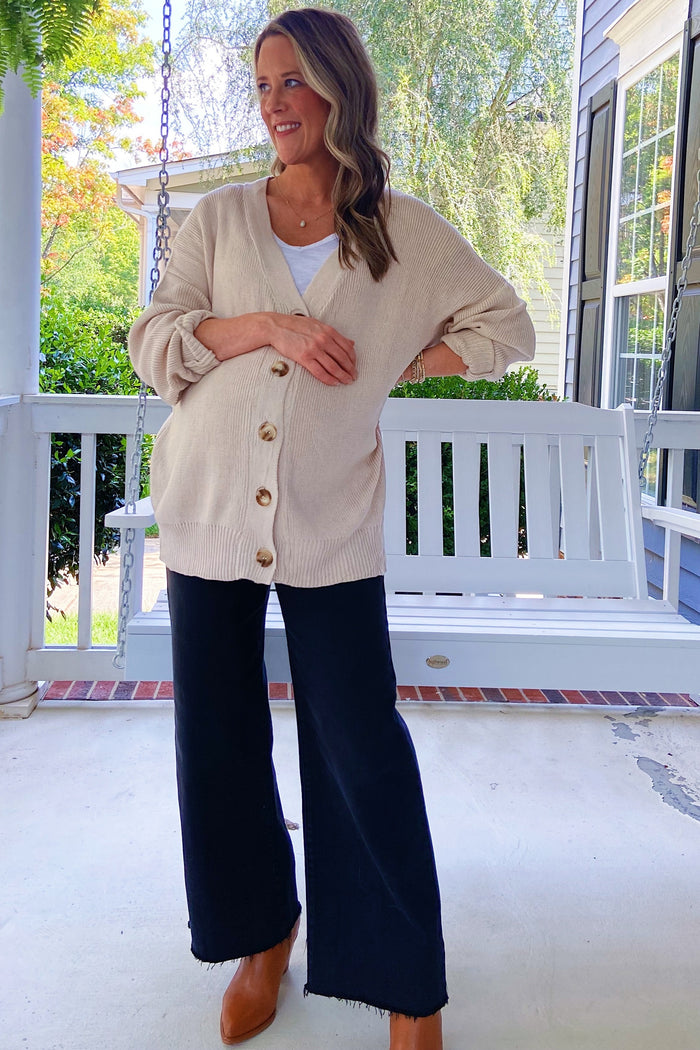 woman wearing a cream maternity cardigan with buttons, white tank top, dark wide-leg pants, and a visible baby bump