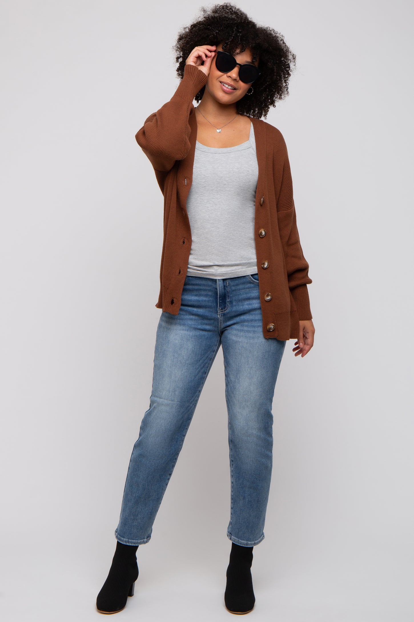 woman wearing a brown button-up cardigan over a grey tank top and blue jeans, accessorized with black sunglasses and ankle boots
