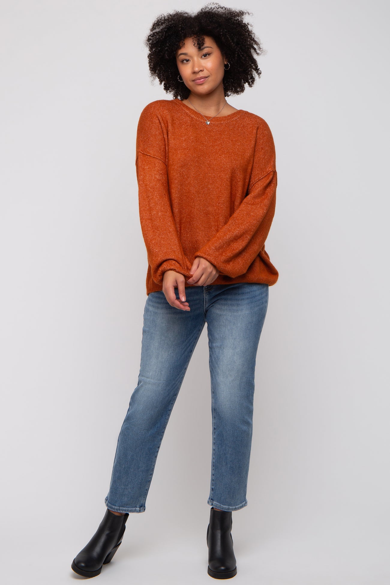 woman wearing an oversized rust orange sweater with long sleeves and blue straight-leg jeans, standing in black ankle boots