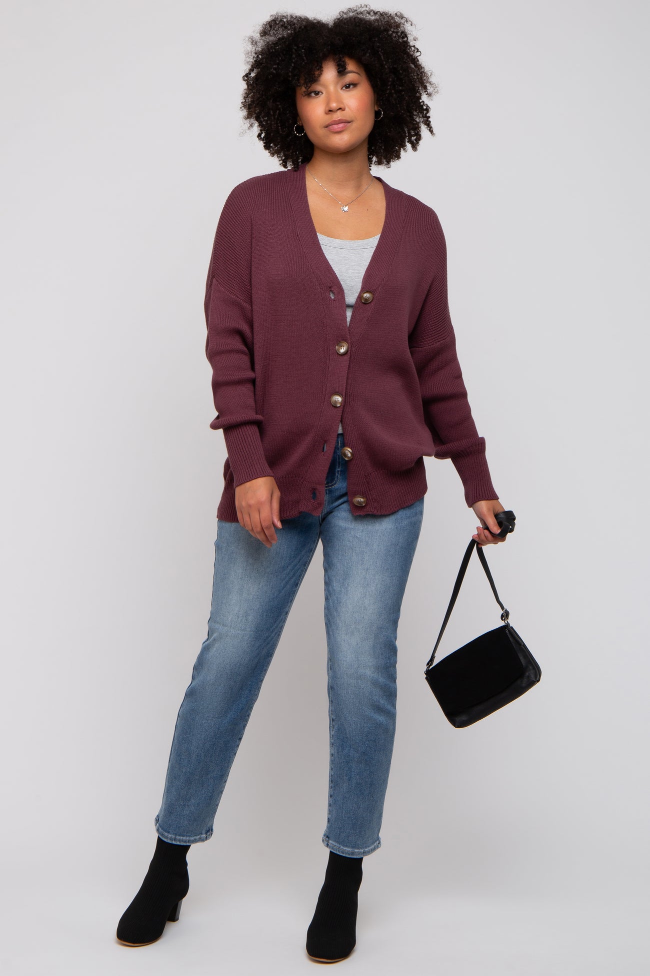 woman wearing a maroon oversized cardigan with buttons, gray top, and blue jeans, carrying a small black purse