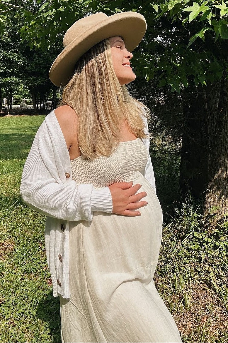 woman wearing a white strapless maternity dress and a cream cardigan, holding her baby bump outdoors
