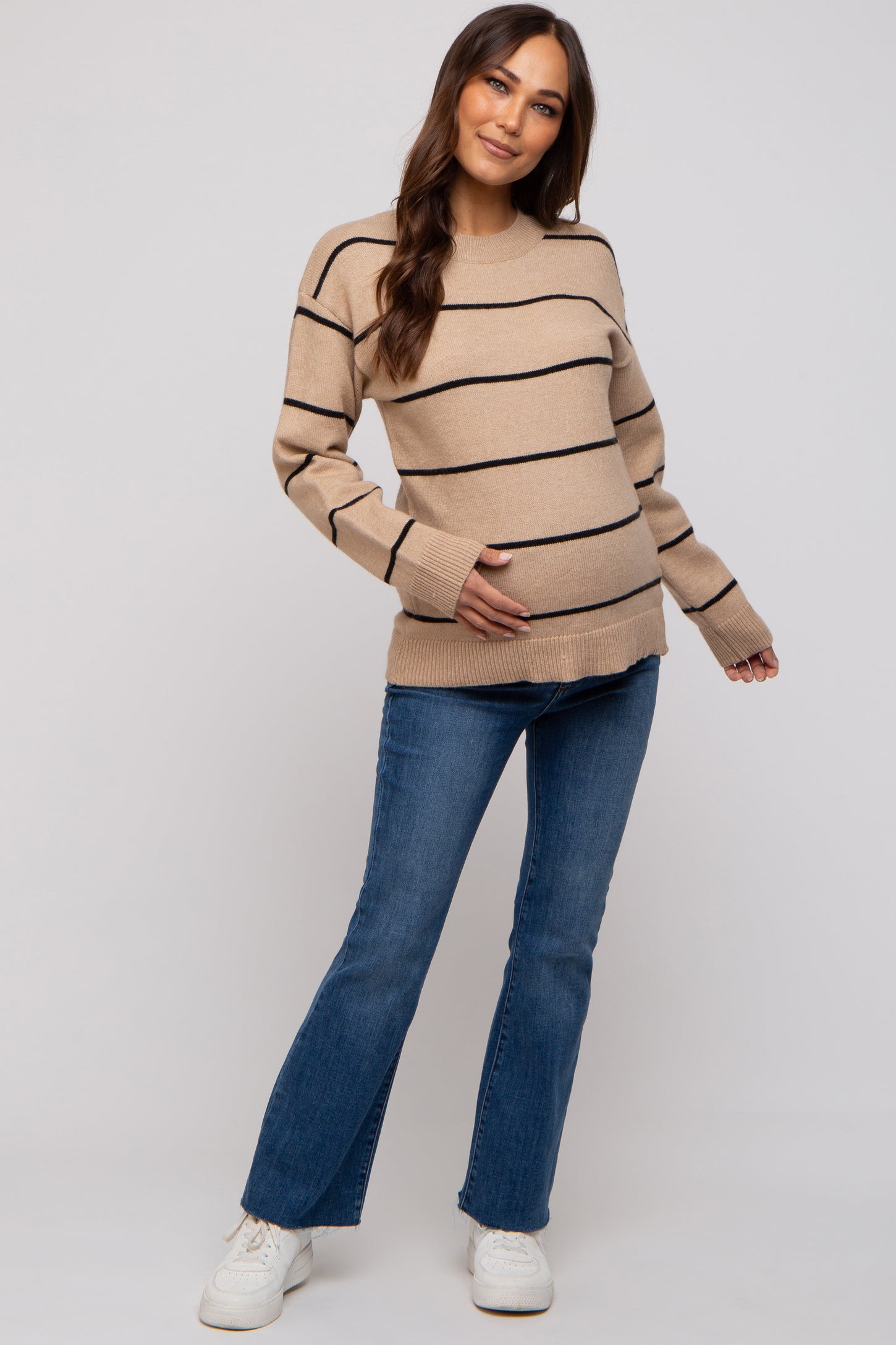 woman wearing a tan striped maternity sweater and blue flared jeans, holding her baby bump