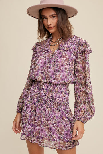 woman wearing a purple floral smocked mini dress with ruffle details and long sleeves, paired with a wide-brimmed hat