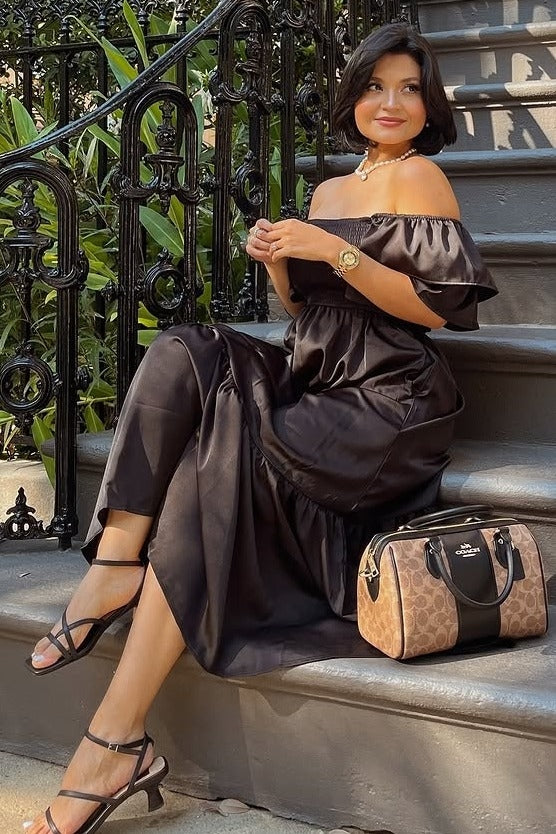 woman sitting on steps wearing a black off-shoulder maxi dress, paired with strappy heels and holding a designer handbag