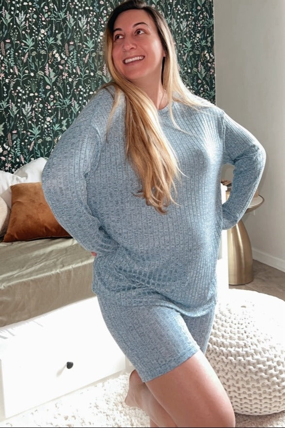 woman wearing a light blue maternity sweater and matching shorts, smiling with hands on hips while showing her baby bump
