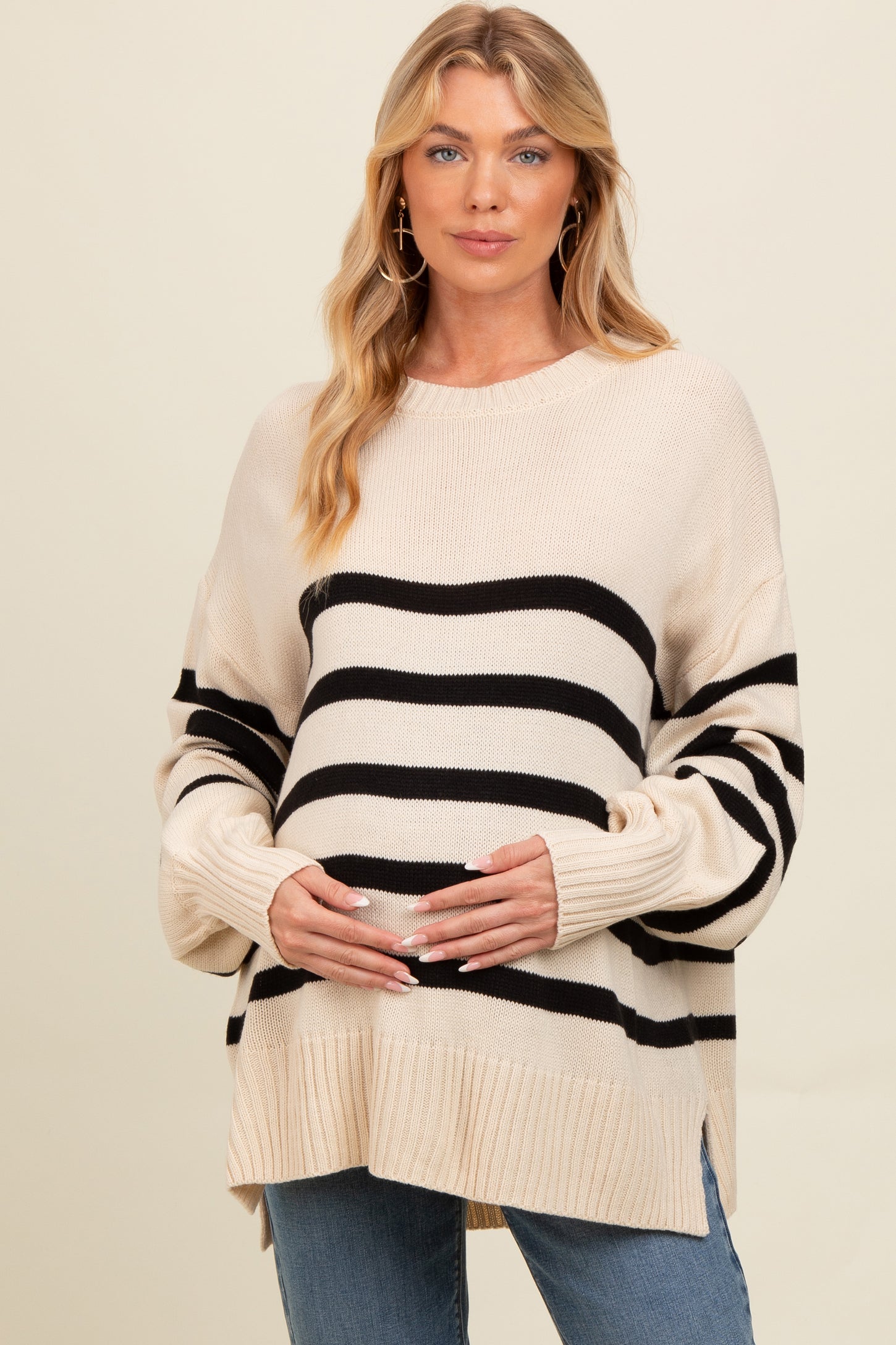 woman wearing a cream and black striped maternity sweater, holding her baby bump, paired with blue jeans