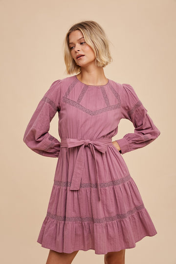 woman wearing a lilac long sleeve dress with lace detailing and a tied waist, styled with a flared hemline