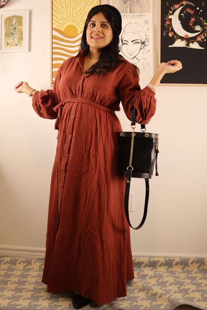 woman wearing a rust-colored long sleeve button-up maxi dress with a belted waist and holding a black handbag